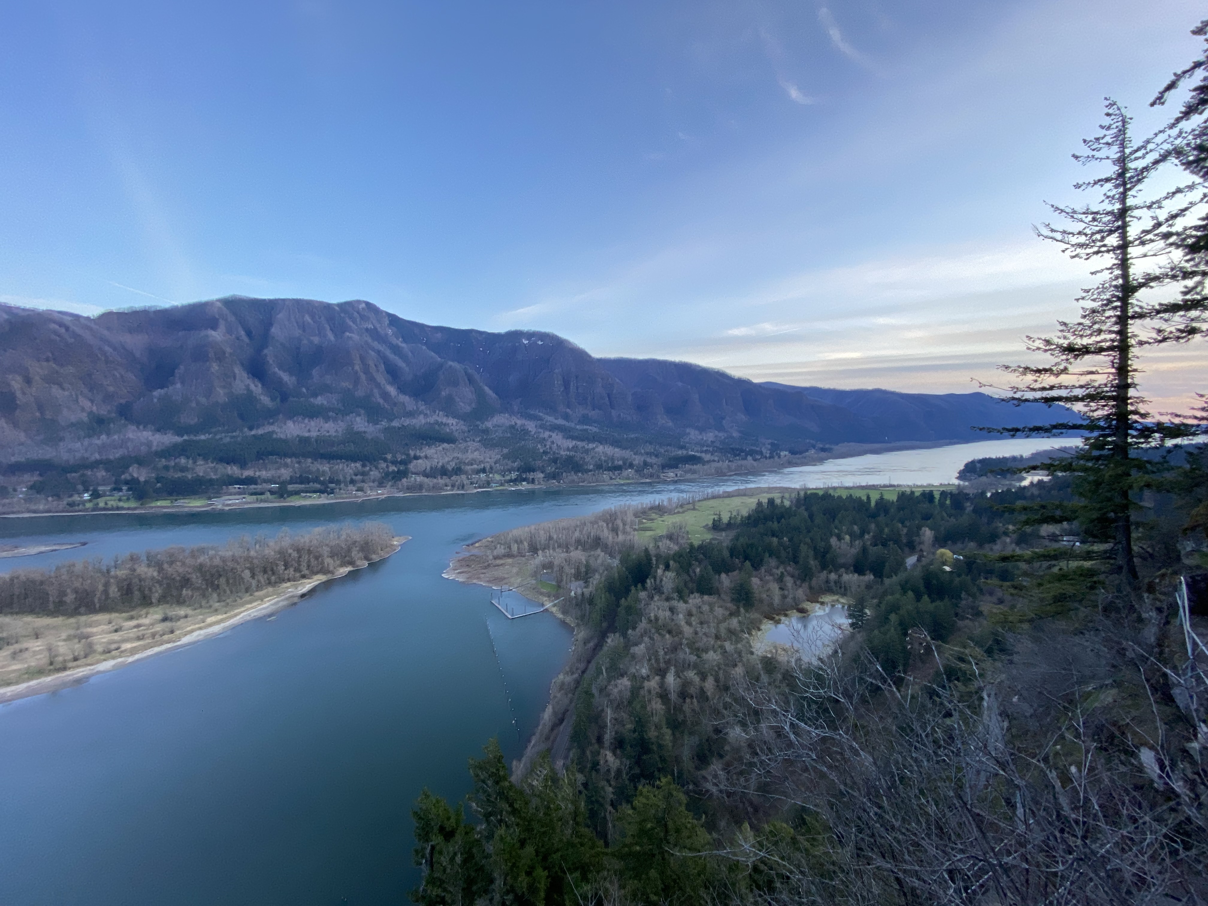 High vantage point photograph of a wide, deep blue river with mountains with evergreen forests in the foreground and mountain cliffs in the background