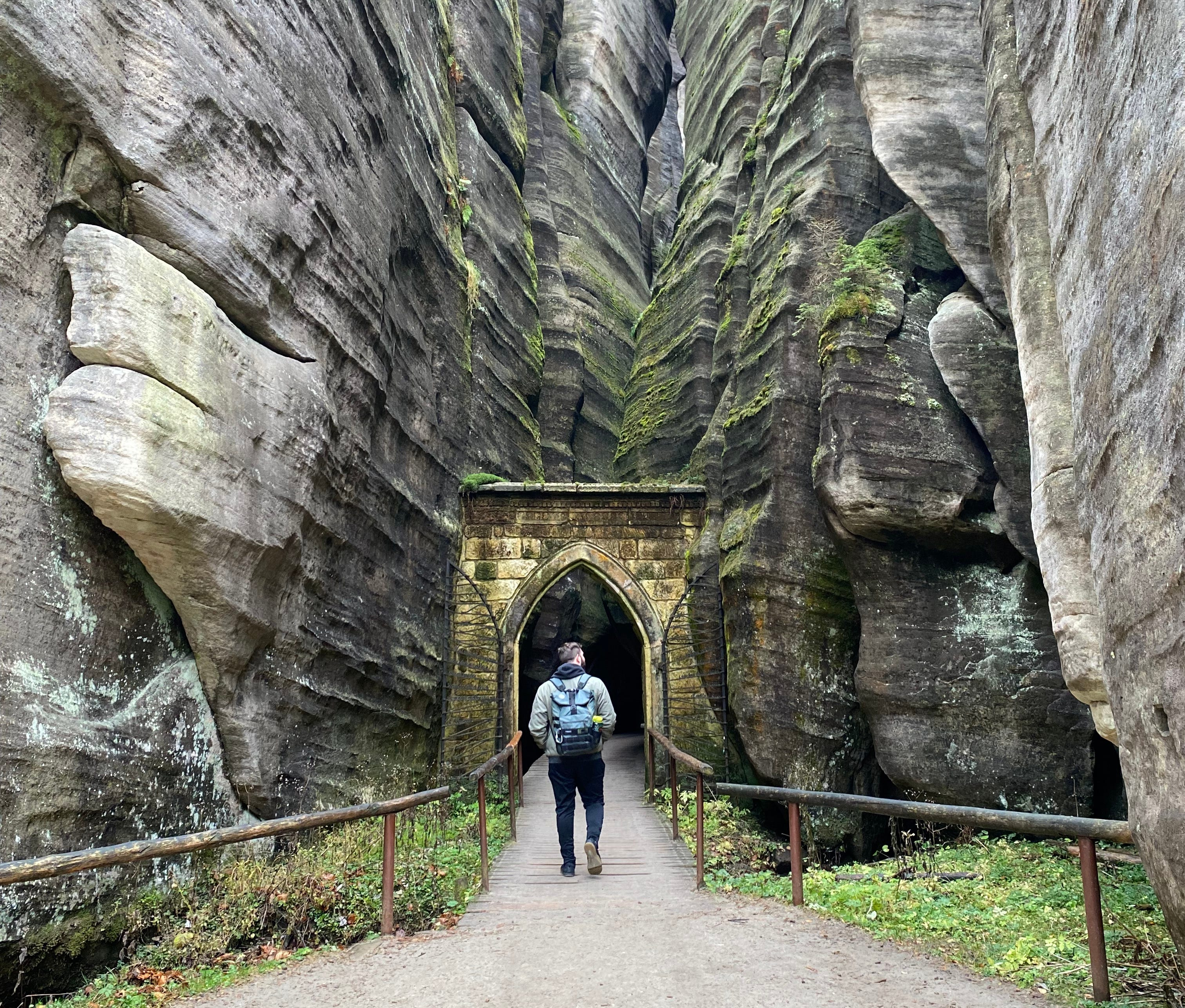 photo of a man with his back turned to the camera walking through a yellow stone arch gate flanked by gray stone slot canyon covered in green moss